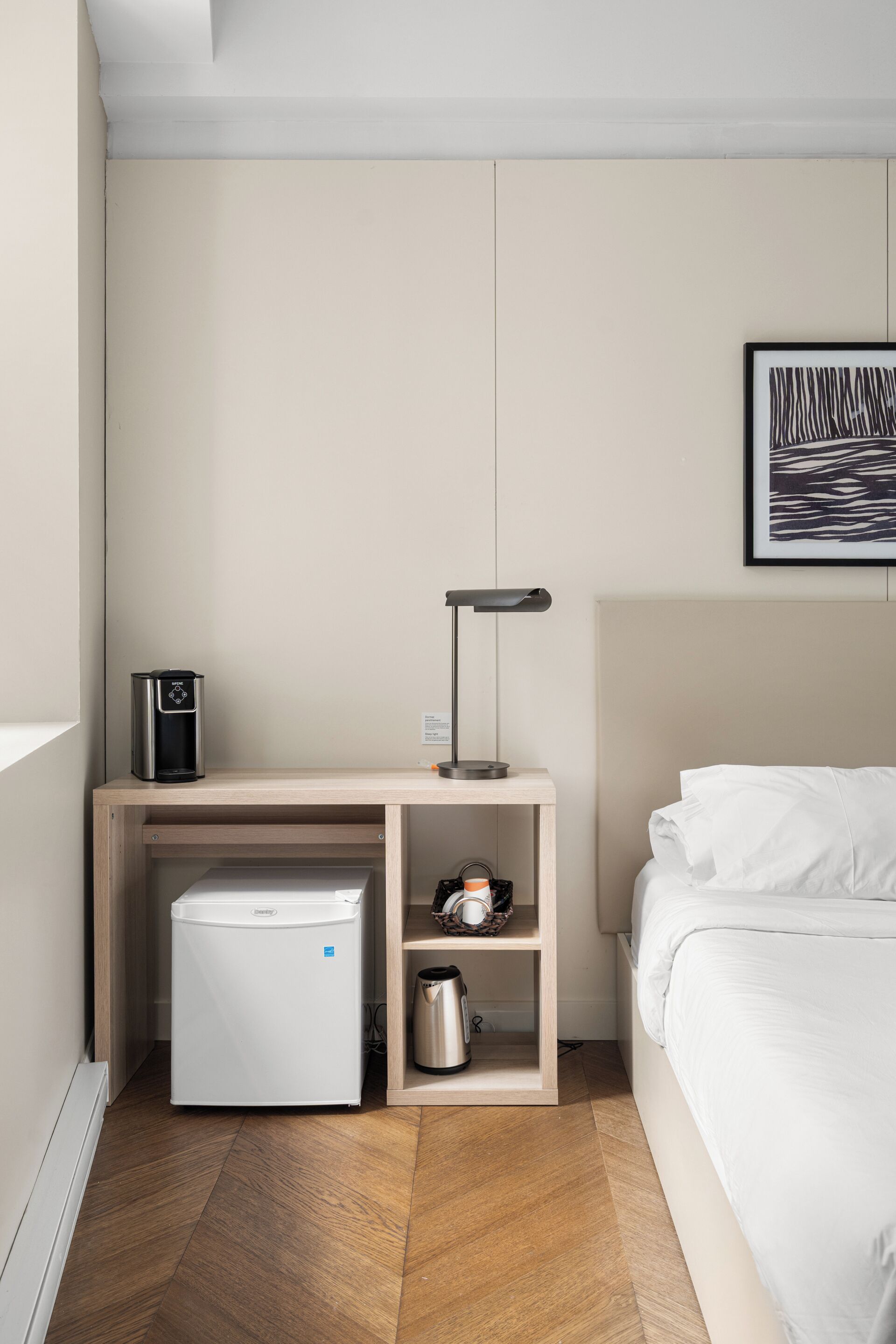 A compact hotel room with a bed, a small desk with a coffee maker, a mini-fridge, a lamp, and a framed artwork on the wall.