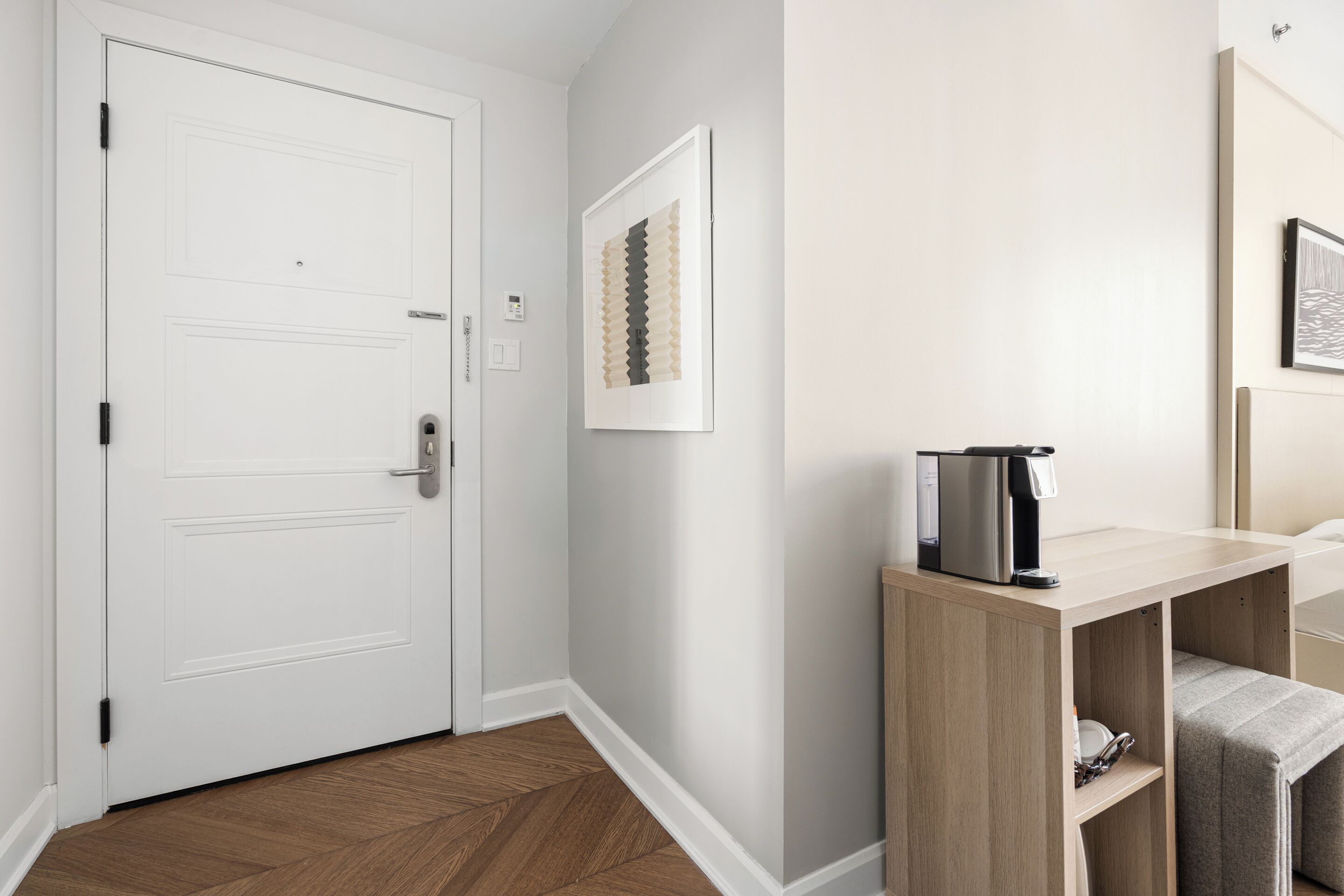 A hotel room with a white door, a coffee machine on a wooden console table, and a framed artwork on the wall.