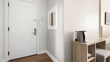 A hotel room with a white door, a coffee machine on a wooden console table, and a framed artwork on the wall.