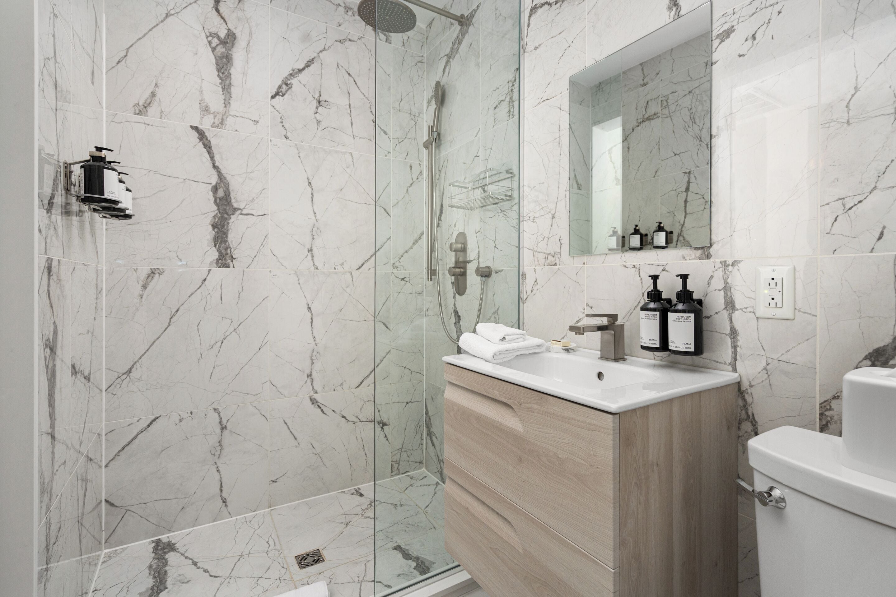 A modern bathroom with a glass-enclosed shower, marble walls, a wooden vanity, and a white sink.