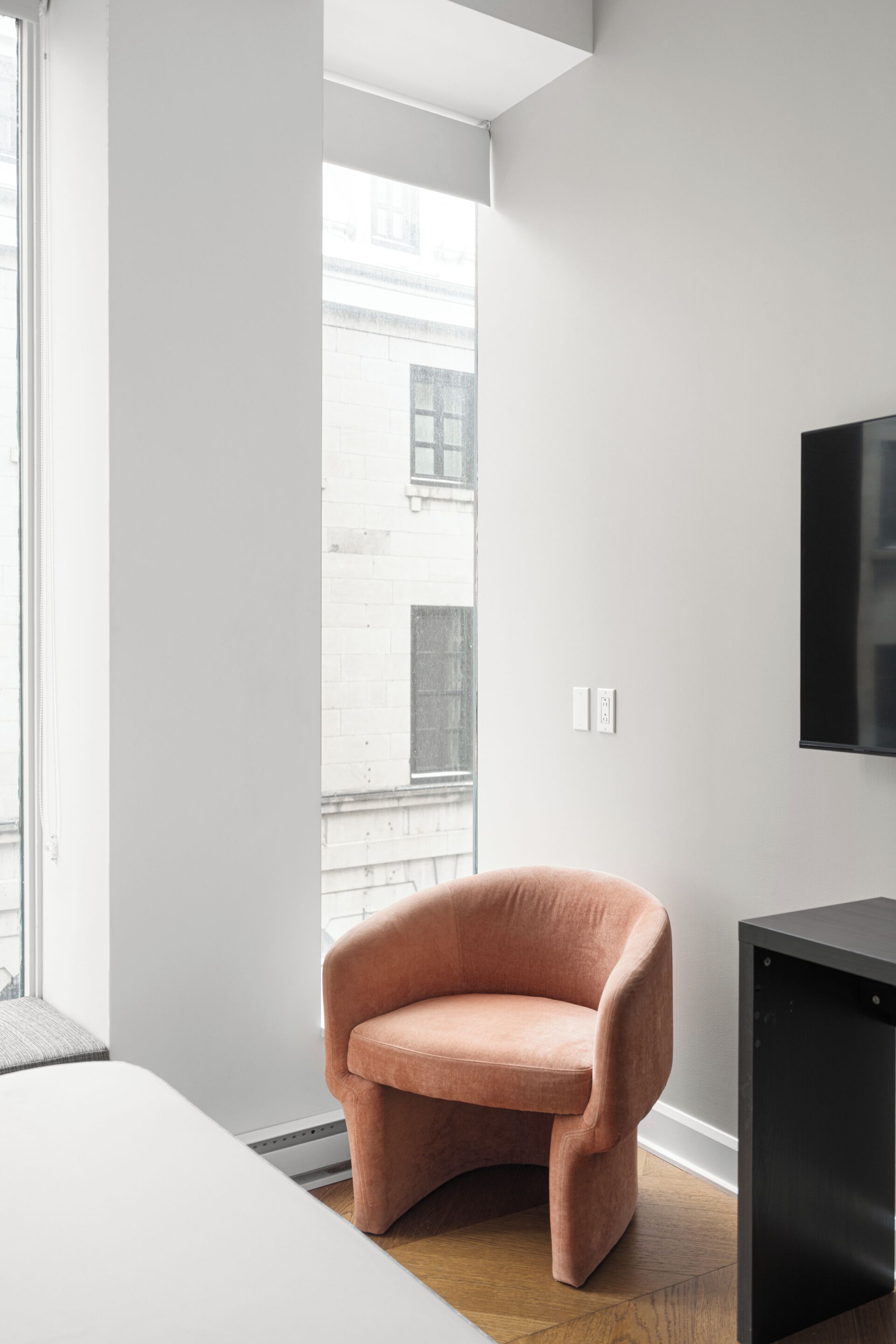 Una habitación moderna con un ventanal grande, un sillón rosa, un aparador negro y una cama blanca.