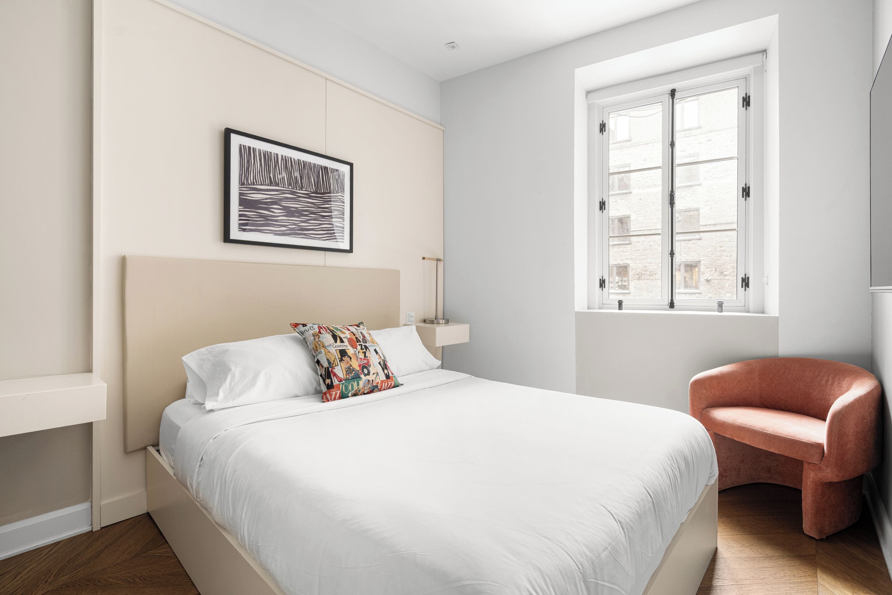 Un dormitorio moderno con una cama grande, un cojín colorido, una obra de arte enmarcada, una ventana con persianas y una silla rosa.