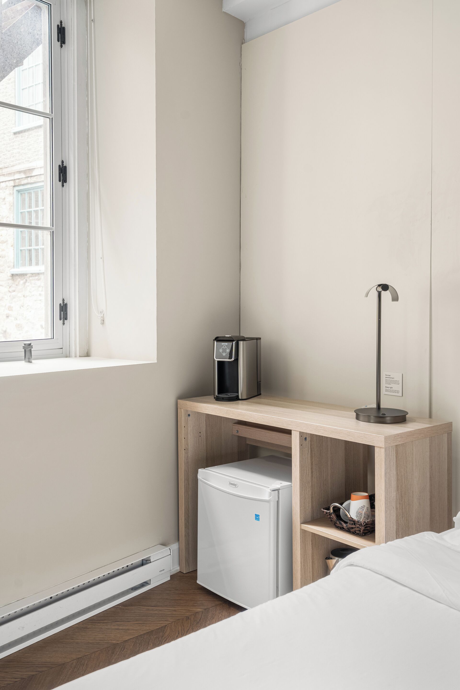 Habitación de hotel con cama, un pequeño escritorio con cafetera, refrigerador y lámpara de noche.