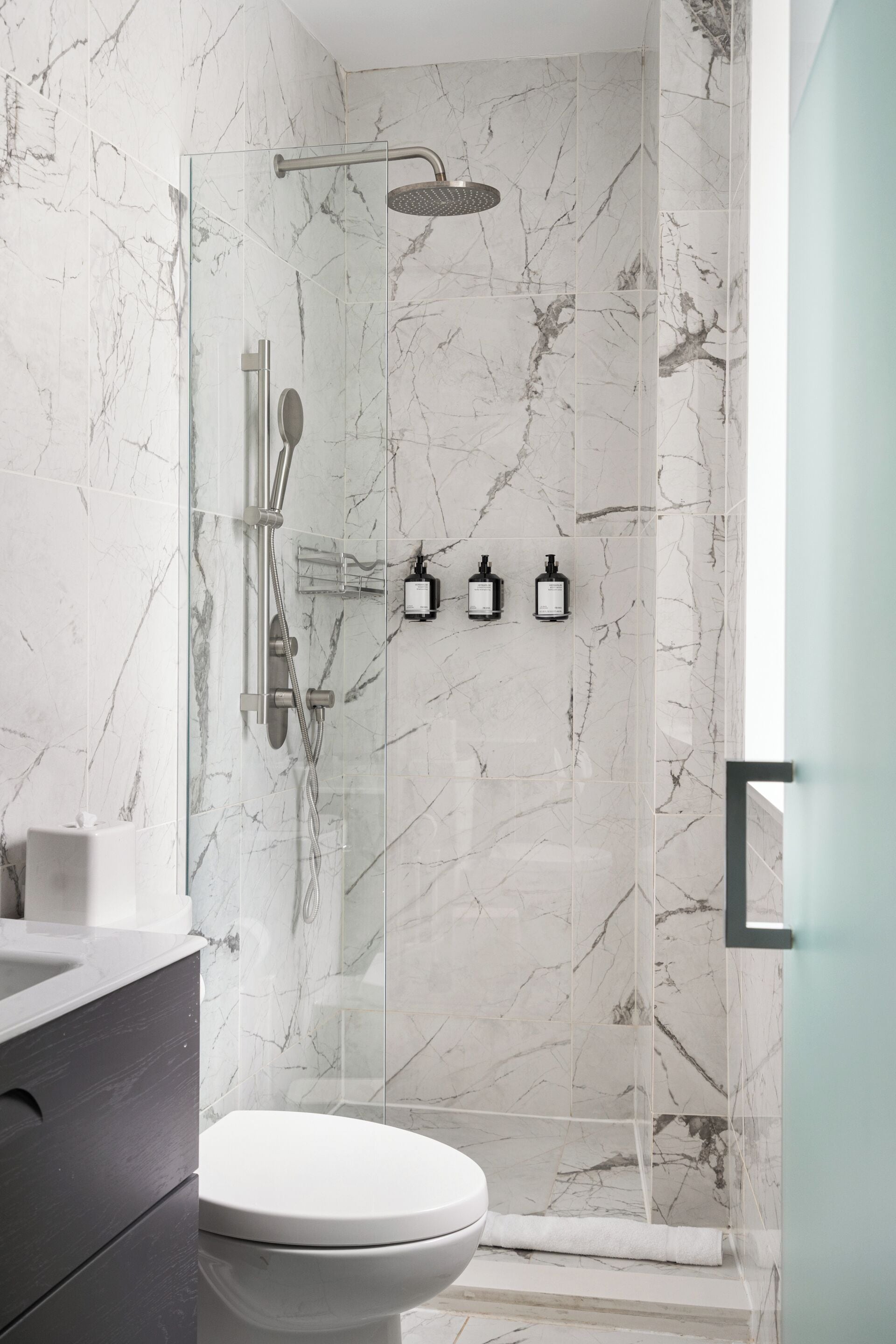 Une salle de bain moderne avec une douche à parois de verre, des murs en marbre, des toilettes blanches et un meuble-lavabo de couleur foncée.