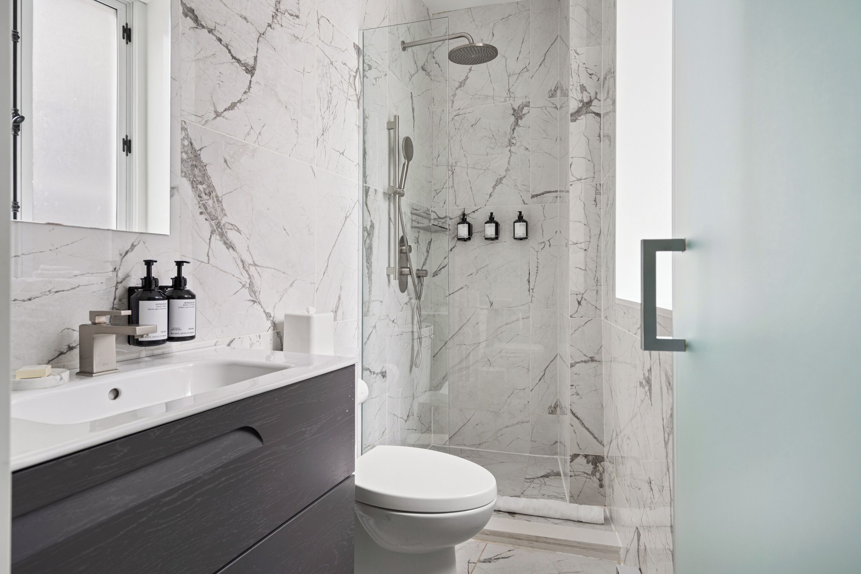 A modern bathroom with a marble shower, a white toilet, and a vanity with soap dispensers.