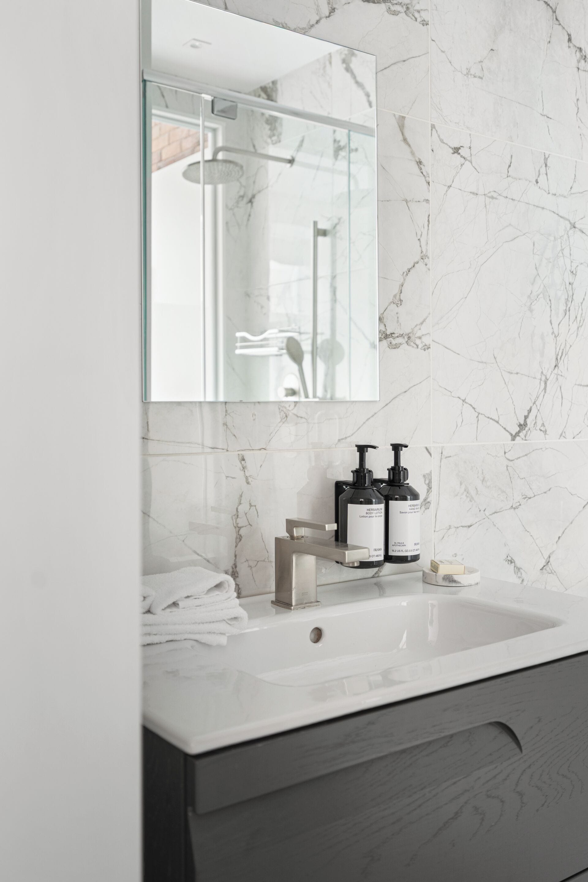 Een moderne badkamer met een marmeren achterwand, een grote spiegel, een witte wastafel en twee zwarte zeepdispensers.