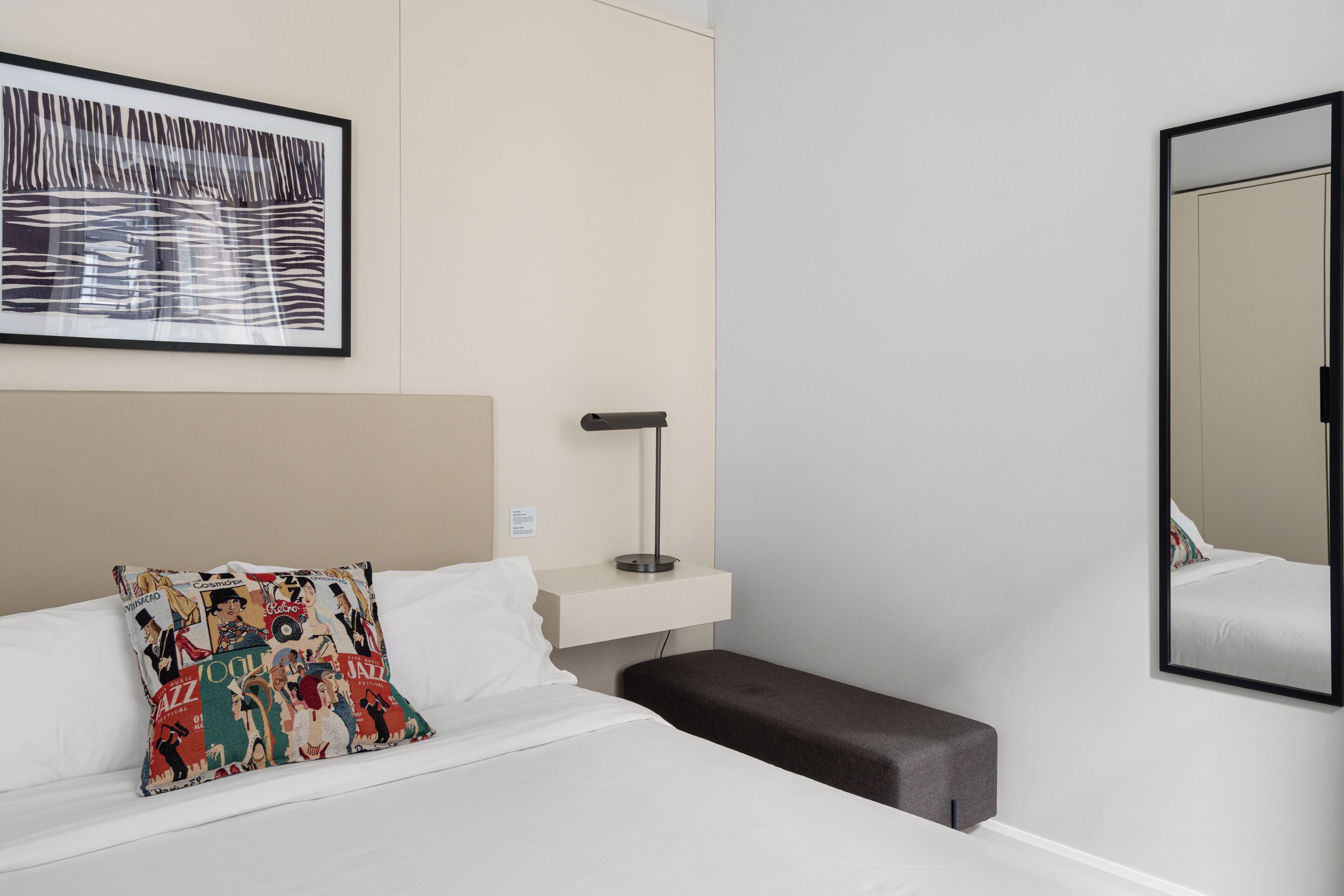 A hotel room with a bed, a bedside table, a lamp, a bench, a framed picture, and a mirror.