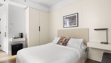Una habitación de hotel moderna con una cama grande, una mesita de noche con lámpara, un escritorio pequeño con cafetera y un armario.