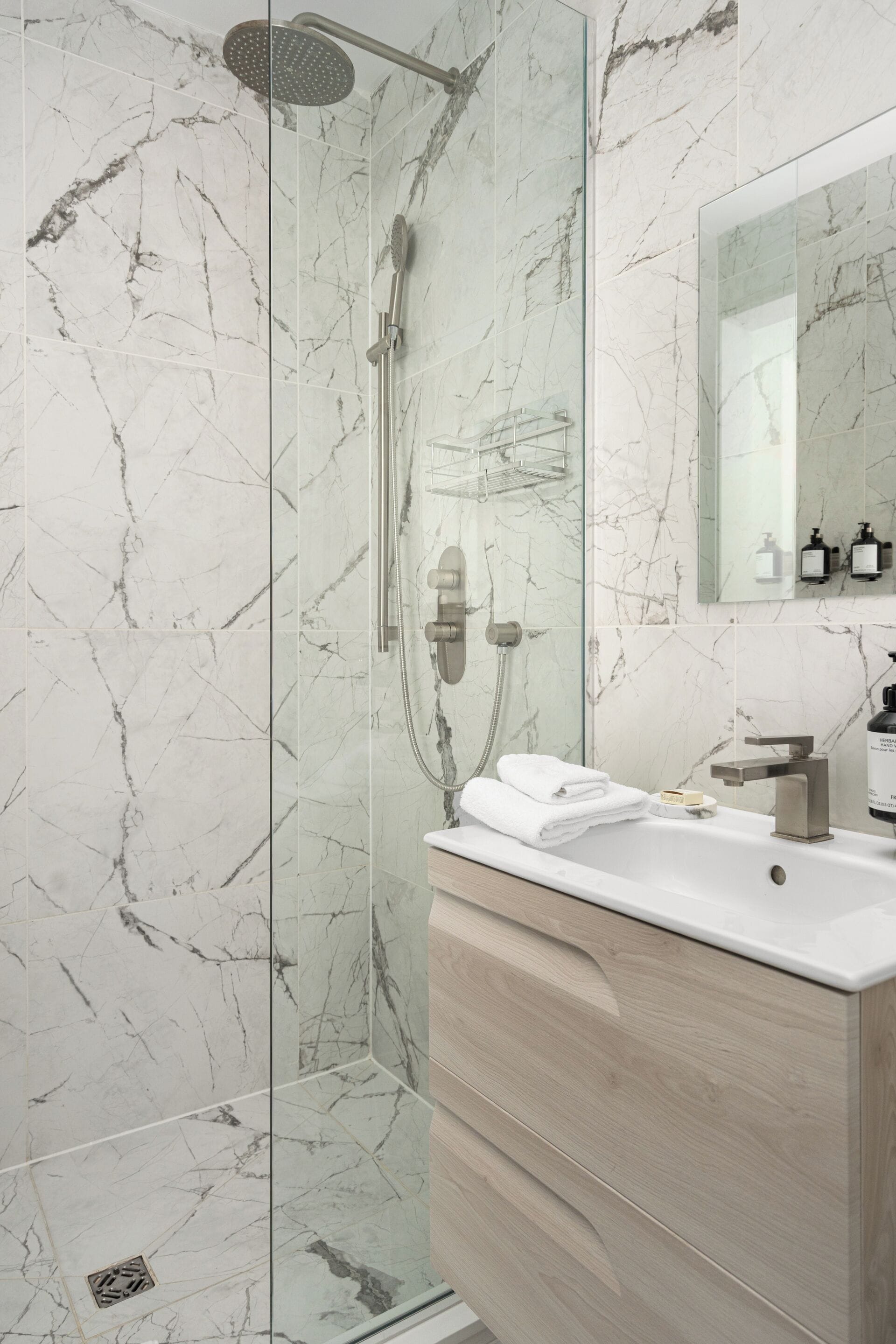 Un baño moderno con ducha envuelta en vidrio, paredes de mármol, tocador de madera y un espejo.
