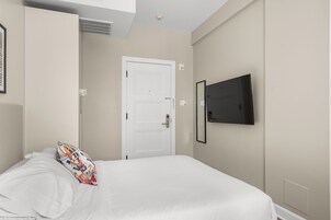 Une chambre à coucher avec un lit, une porte blanche, un téléviseur fixé au mur et une grille d’aération au-dessus de la porte.