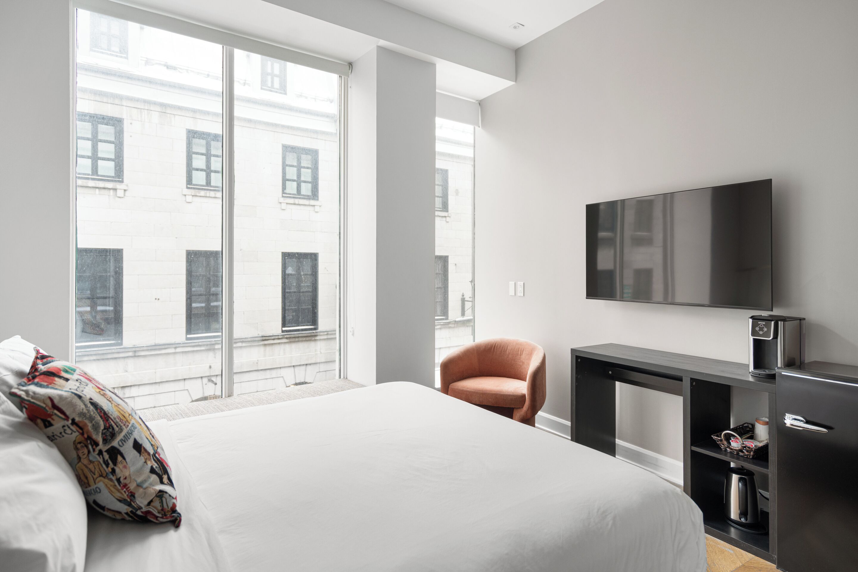 A hotel room with a large bed, a TV mounted on the wall, a small desk with a kettle, and a window with a view of buildings.