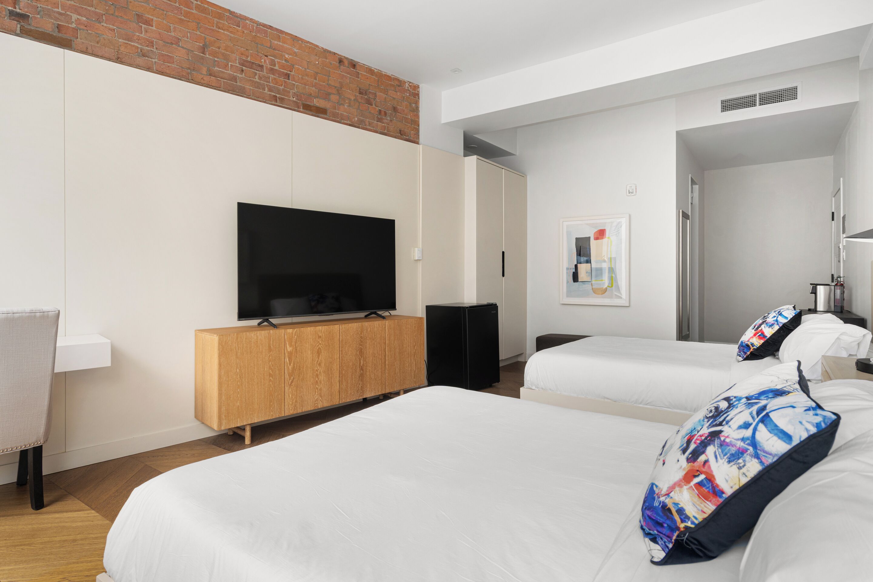 A hotel room with a TV on a wooden cabinet, two beds with white bedding, a brick wall, and a door leading to another room.