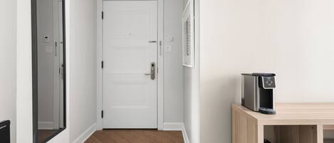 A hallway with a white door, a coffee machine on a wooden shelf, and a framed picture on the wall.