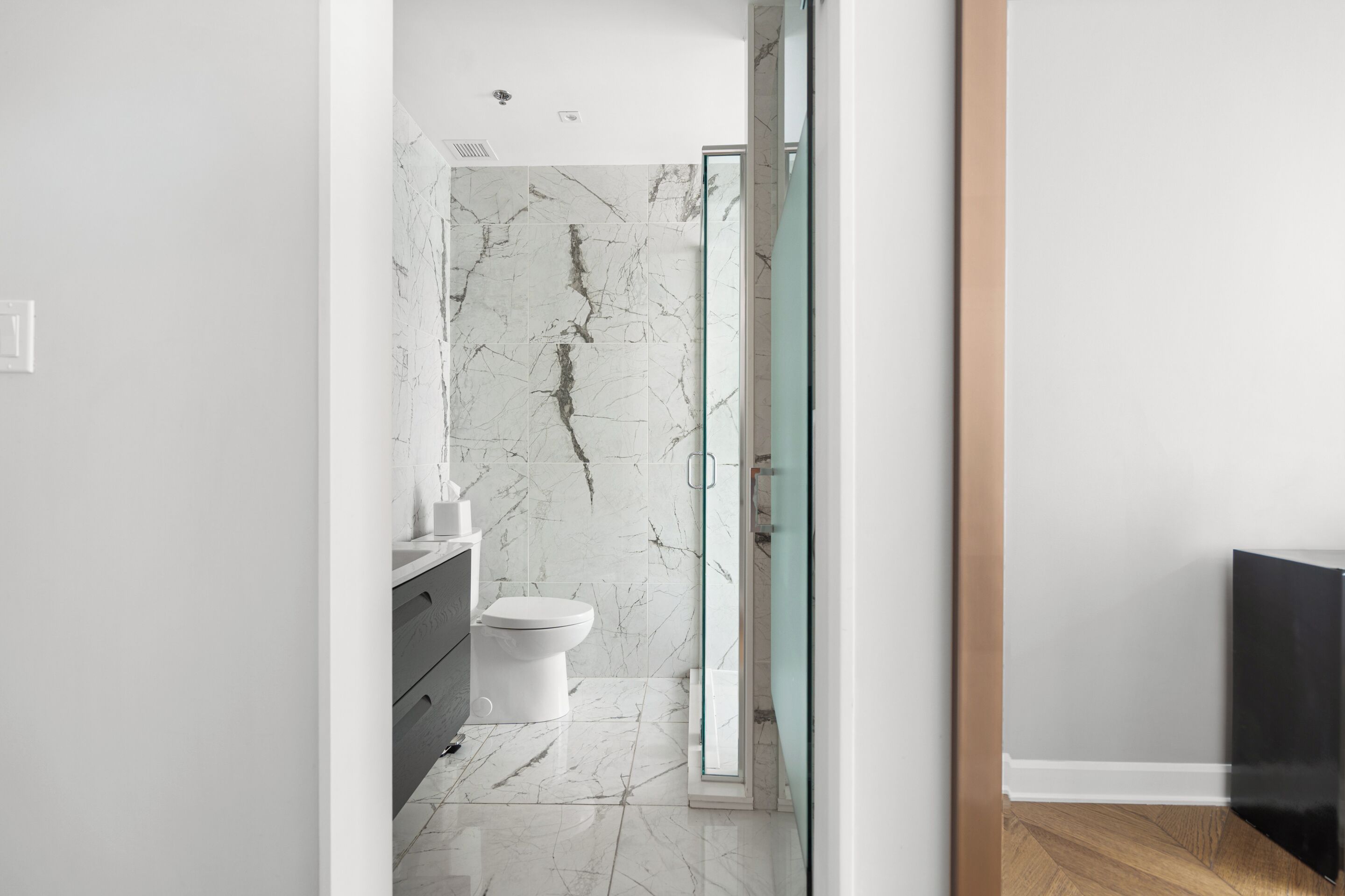 A modern bathroom with a marble wall, a glass shower enclosure, and a wooden floor.
