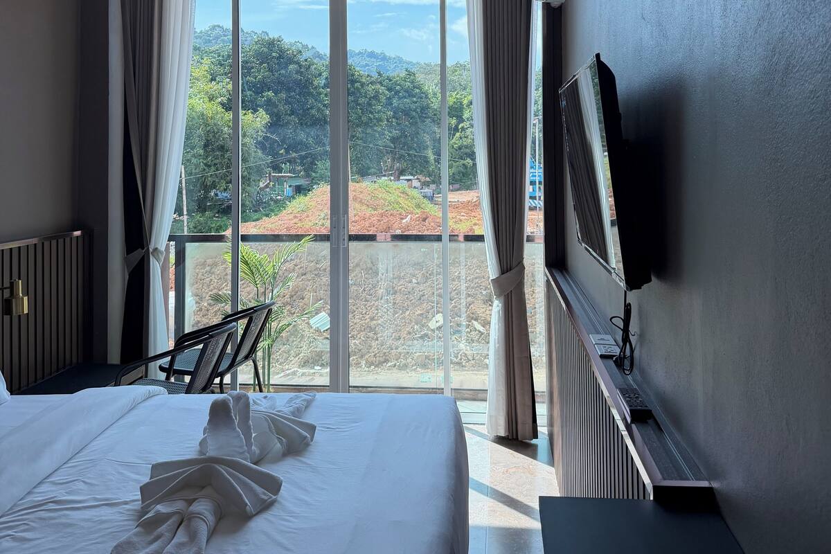 Ein Hotelzimmer mit einem Bett, einem Deckenventilator, einem großen Fenster mit Vorhängen, einem an der Wand befestigten Fernseher und Blick auf Grünflächen.