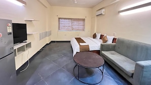 A hotel room with a bed, a sofa, a small table, a TV, and a window with blinds. - GoBravo Residences – Kapashera, New Delhi (New Delhi)