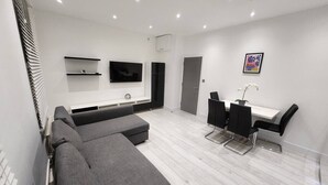 A modern living room with a grey sectional sofa, a dining area with a white table and black chairs, a flat-screen TV mounted on the wall, and a potted plant on the dining table. - Modern 2BR London Apt Near Tube (Greater London)