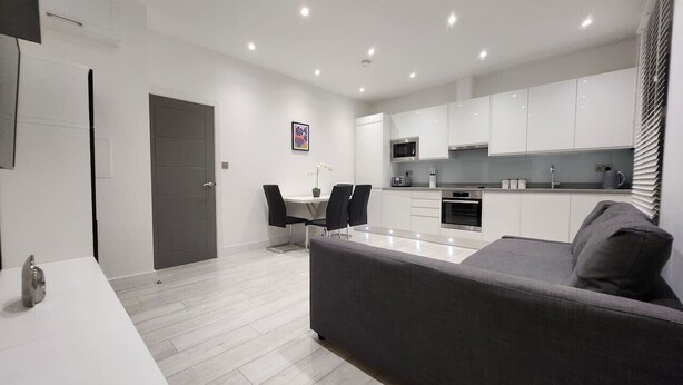 A modern kitchen with white cabinets, a dining area with a table and chairs, and a grey sofa. - Modern 2BR London Apt Near Tube (Greater London)