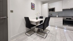 A modern kitchen with white cabinets, a stainless steel microwave, and an oven. There is a dining table with four chairs and a vase of flowers. - Modern 2BR London Apt Near Tube (Greater London)