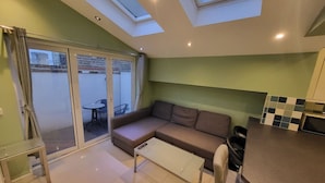 A modern living room with a sofa, a coffee table, and a kitchen area with a counter and cabinets. There is a sliding glass door leading to an outdoor area with a table and chairs. - Large 4 Bedroom House In Central London (Greater London)