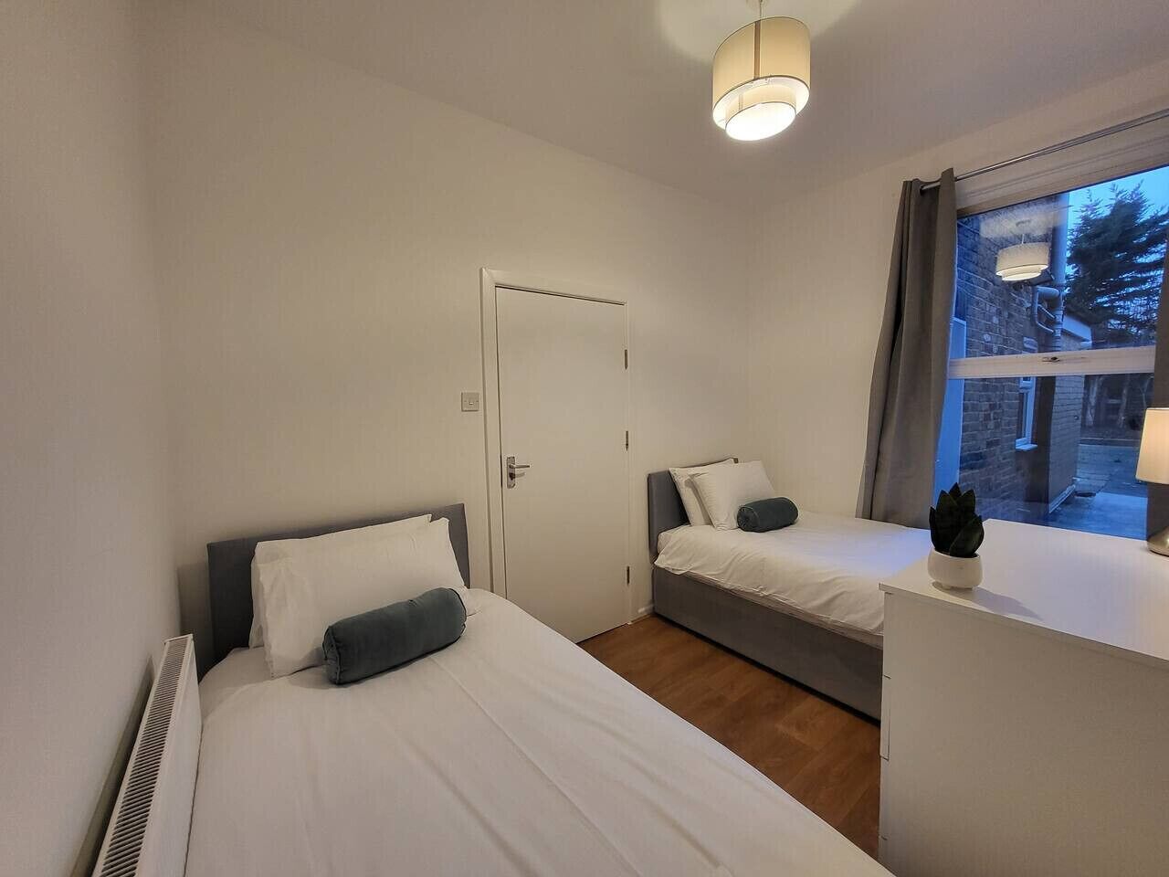 A bedroom with two beds, a desk, and a window with a view of trees and buildings.