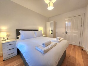 A neatly made bed with white linens, a bedside table with a lamp, and a wardrobe in a hotel room. - Spacious 2-Bed Flat - Close to Tube, Sleeps 6 (Greater London)