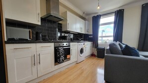 A modern kitchen with white cabinets, a black countertop, and a built-in oven. There is a grey sofa and a window with curtains in the living area. - Tooting Bec Central Apartment by London Tube (Tooting Bec, London)