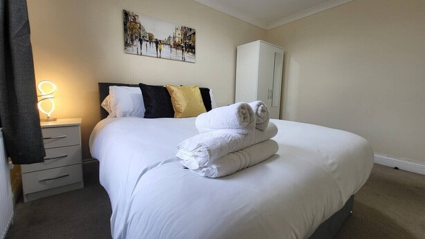 A neatly made bed with white linens and rolled towels, a bedside lamp, a nightstand, a wardrobe, and a wall-mounted picture. - Tooting Bec Central Apartment by London Tube (Tooting Bec, London)