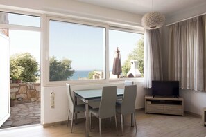 Een kamer met een eettafel en stoelen, een televisie op een dressoir, en grote ramen met uitzicht op zee.