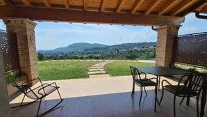 Une terrasse couverte avec une table et des chaises, un chemin de pierres menant à une pelouse, et une vue sur des collines lointaines.
