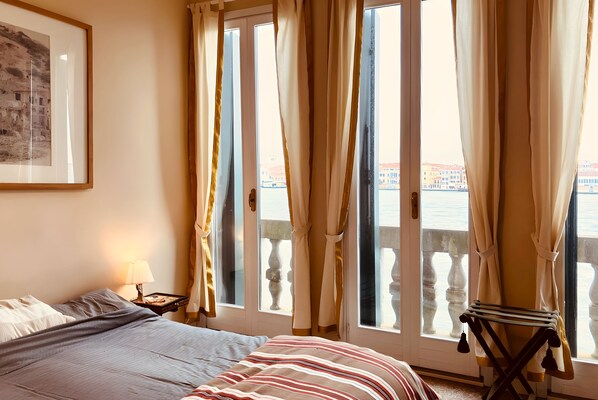 Een hotelkamer met een bed, een nachtkastje met een lamp, een gepresenteerd schilderij aan de muur en uitzicht op een gebouw door het raam.
