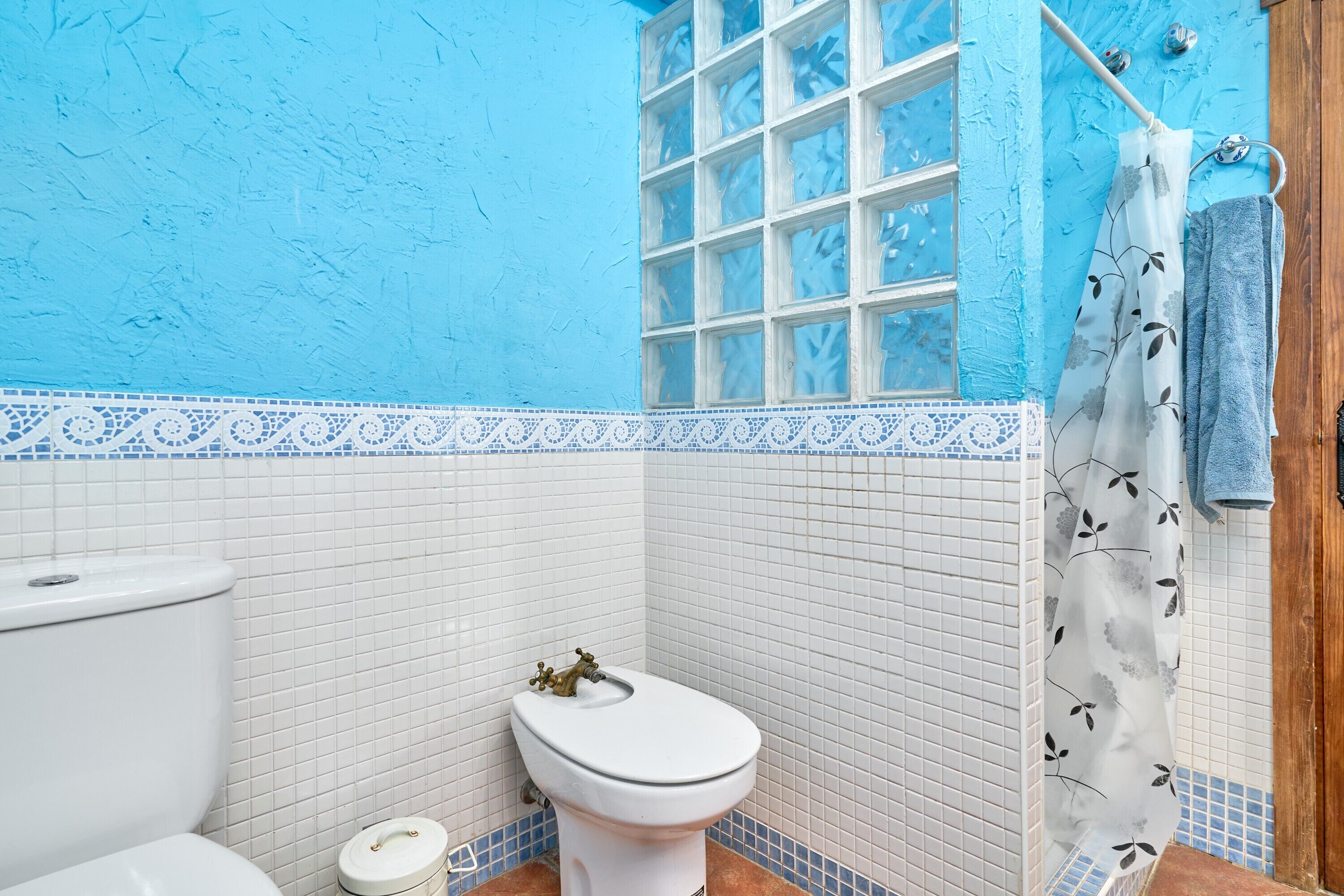 Een badkamer met een wit toilet, een wastafel, een douchegedeelte betegeld met blauwe tegels en een douchegordijn met dessin.