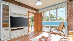 Ein Wohnzimmer mit Fernseher, Holzschränken, einem Korbstuhl und Blick durch ein Fenster.
