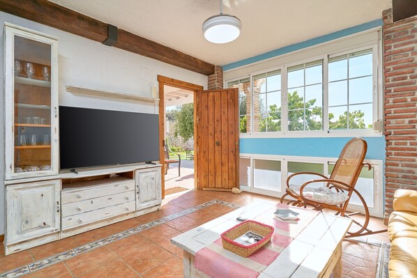 Un salon avec un téléviseur, des meubles en bois, un fauteuil en osier et une vue par la fenêtre.