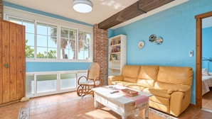 Ein Wohnzimmer mit einer Couch, einem Sessel, einem Couchtisch, einem Bücherregal und einem Fenster mit Blick auf Palmen.