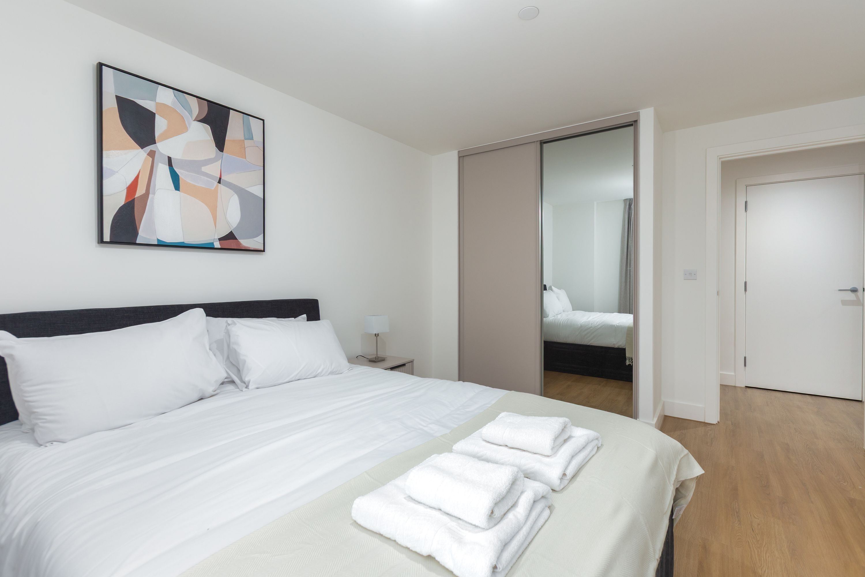 Ein modernes Hotelzimmer mit einem ordentlich bezogenen Bett, einem Nachttisch mit Lampe und einem großen abstrakten Gemälde an der Wand.
