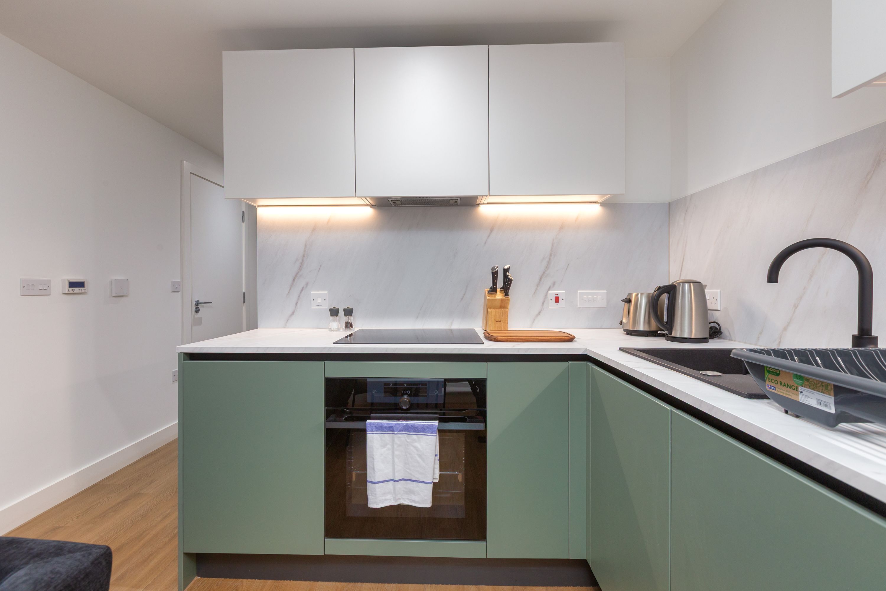 Een moderne keuken met een inbouwoven, een spoelbak en een aanrecht met een waterkoker en een snijplank.