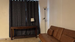 Een woonkamer met een bruine bank, een houten console en een lamp, afgedekt met een donker gordijn.