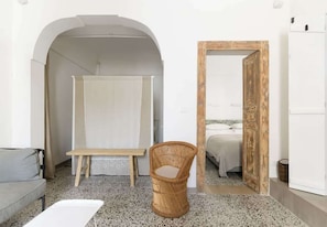 A room with a grey sofa, a wicker chair, a wooden bench, and a bed with a white headboard. - Terrazza Costa - The Fab Stay (Ostuni)
