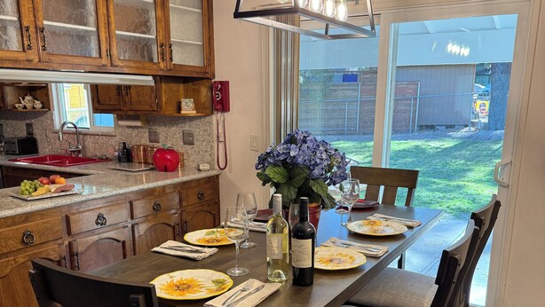 Comedor con una mesa de madera dispuesta para cuatro personas, rodeada de sillas de madera oscura. Un ventanal ofrece vista a un jardín trasero.
