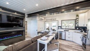 Una cocina moderna con armarios blancos, un comedor con una mesa blanca y cojines marrones, y una chimenea con llama azul.