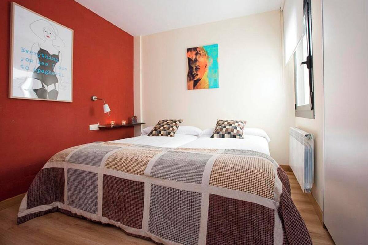 A bedroom with a red accent wall, a bed with patterned bedding, a bedside table with a lamp, and a window with a view.