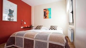 Ein Schlafzimmer mit roter Akzentwand, einem Bett mit gemusterten Betttextilien, einem Nachttisch mit Lampe und einem Fenster mit Blick.