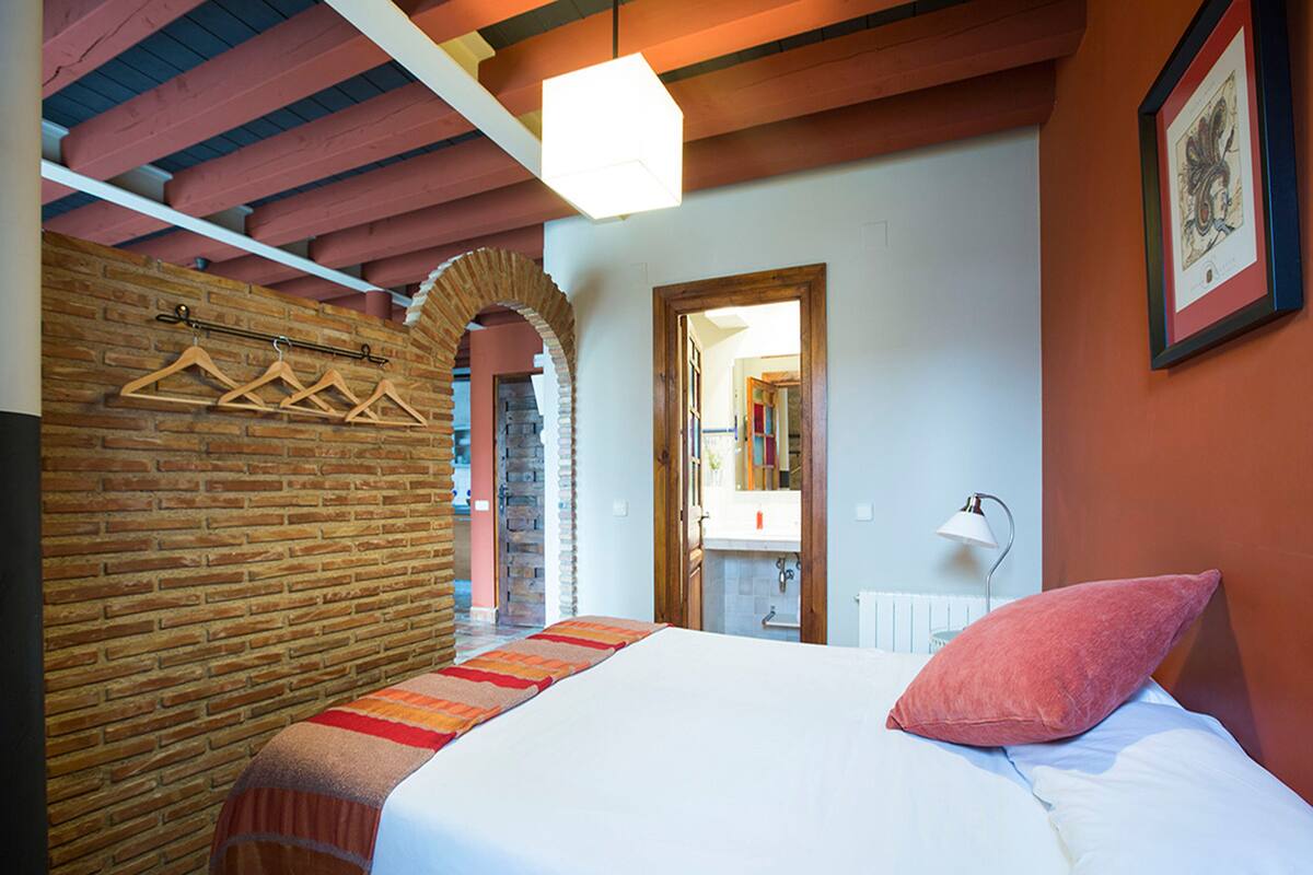 A bedroom with a bed, a brick wall, wooden beams, a hanging light fixture, and a framed picture on the wall.