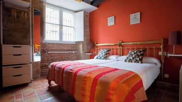 Een slaapkamer met een bakstenen muur, een bed met een gestreepte deken en een raam met witte luiken.
