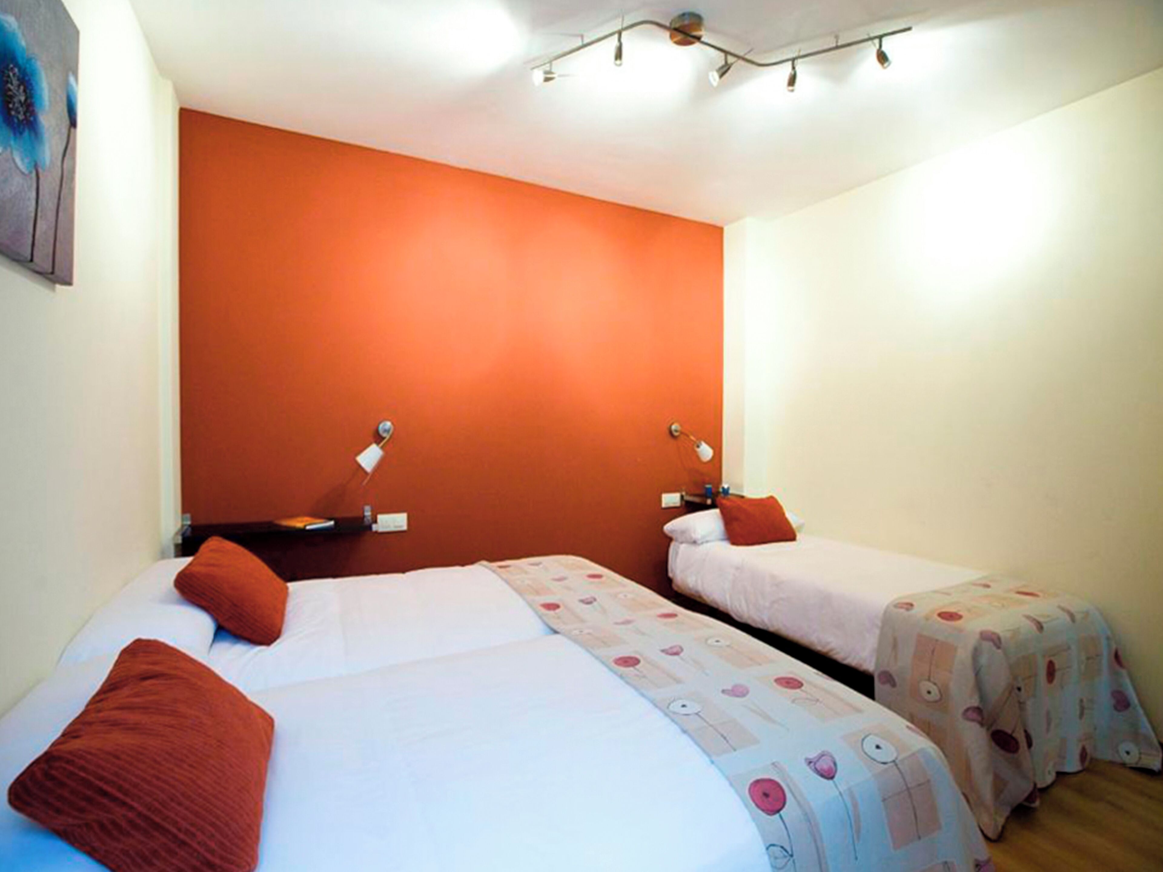 Ein Hotelzimmer mit zwei Betten, jedes mit einem roten Kissen und einer gemusterten Bettdecke. Zwei Nachttische mit Lampen stehen bereit, und die Wände sind orange gestrichen.