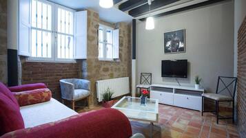 A cozy living room with a sofa, armchair, and a coffee table. There is a TV on a white cabinet, a window with bars, and a stone wall.