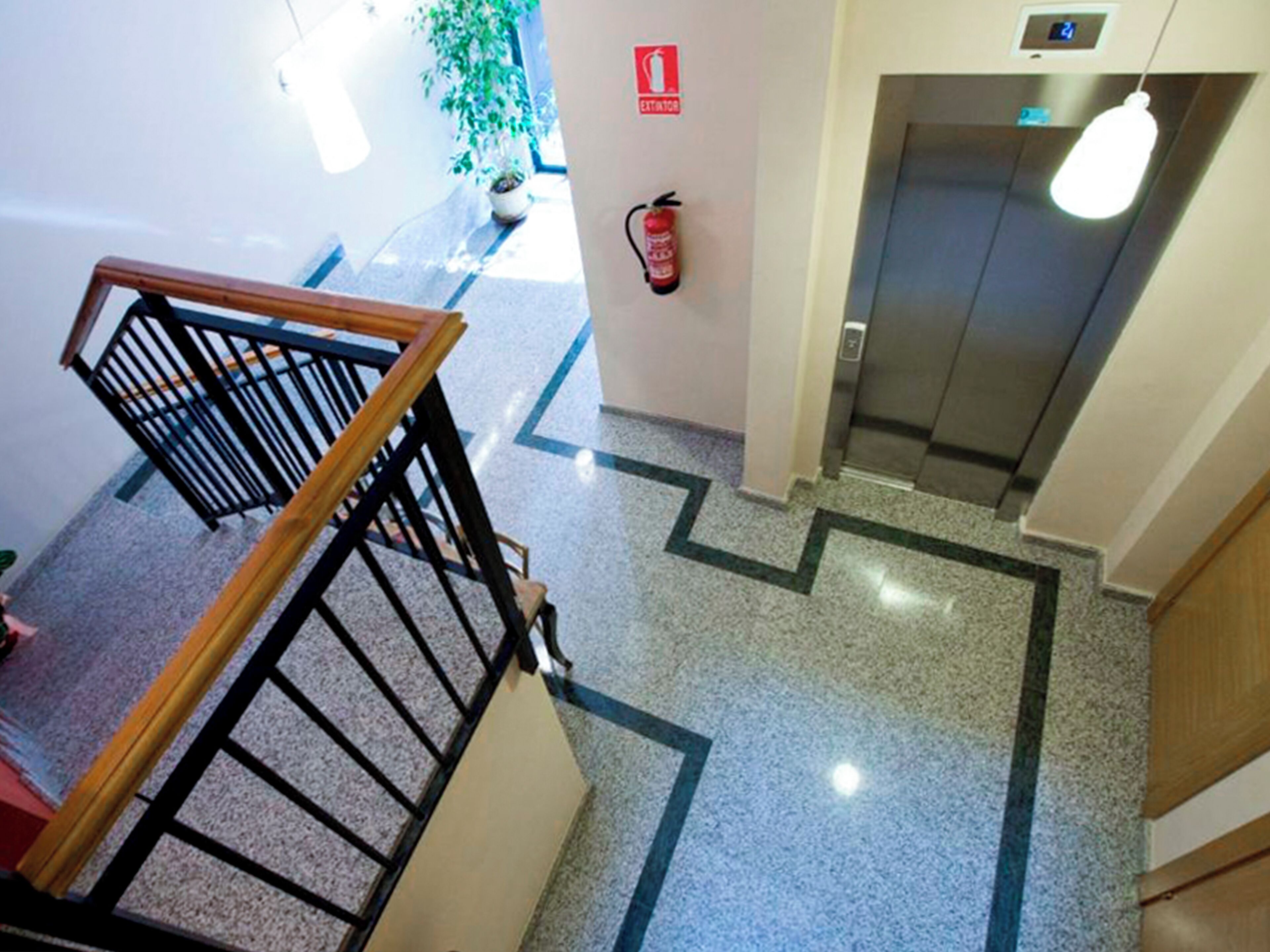 A hotel lobby with a wooden staircase, a fire extinguisher, an elevator, and a potted plant.