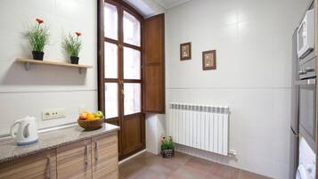 Uma cozinha com armários de madeira, bancada com chaleira e fruteira, janela com vista, radiador e máquina de lavar.