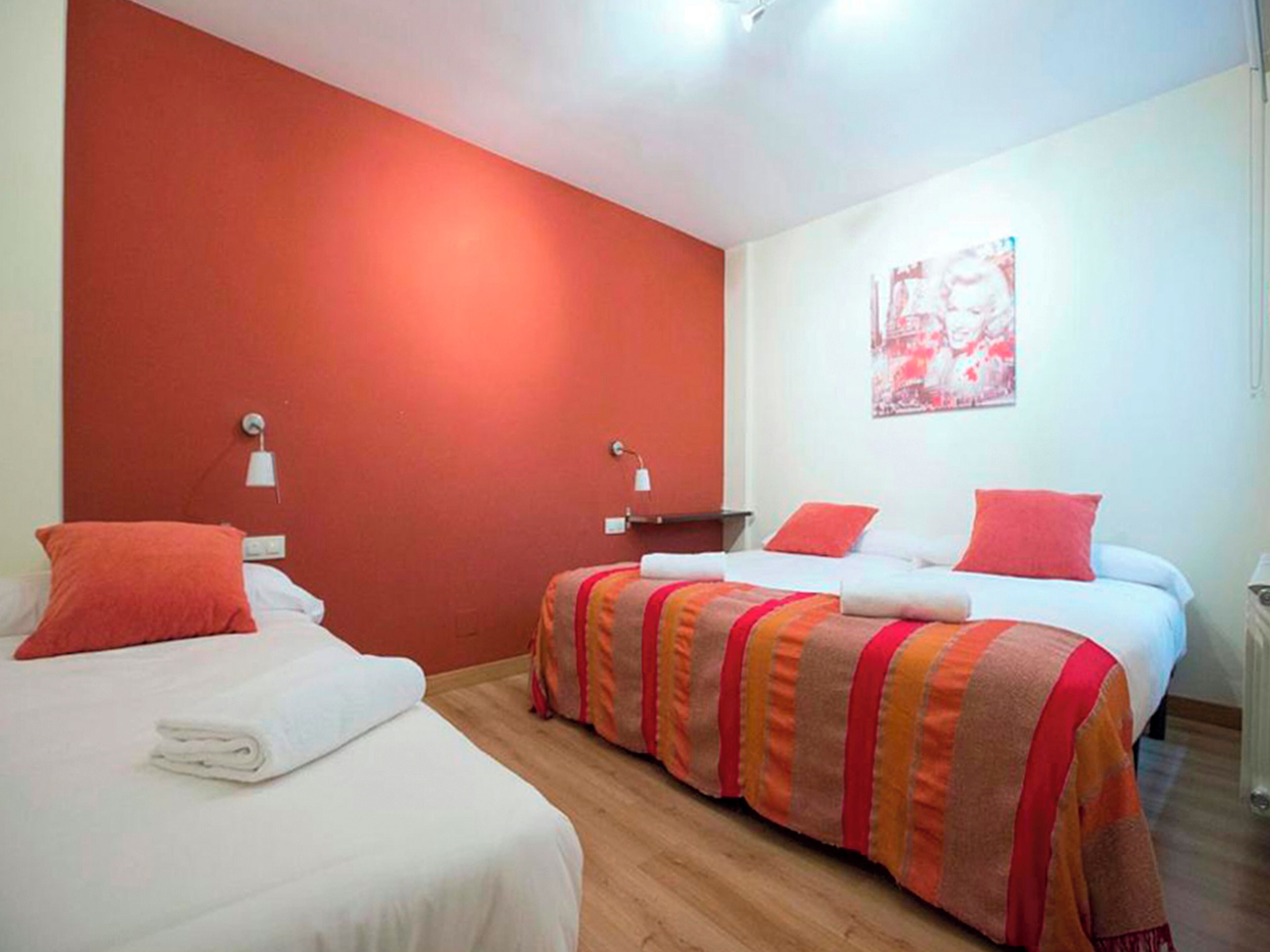 A hotel room with two beds, each with a red pillow and a white blanket. There are two bedside tables with lamps, and a painting on the wall.